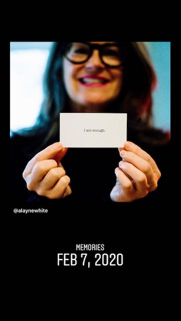 Woman with long dark hair and bold glasses, smiling, holding a small card with the words "I am enough".
