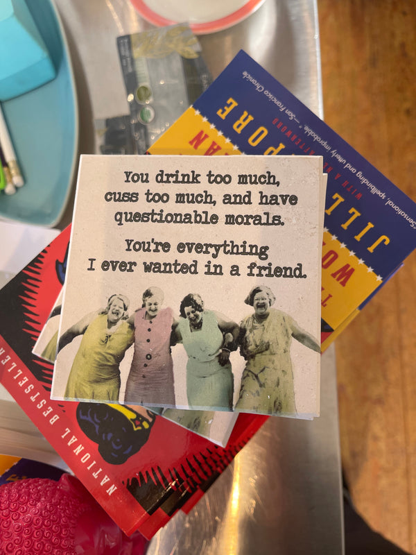 Blurred background with a square drink coaster in focus with text "You drink too much, cuss too much, and have questionable morals. You're everything I ever wanted in a friend. Picture of four woman linked arms and laughing below.