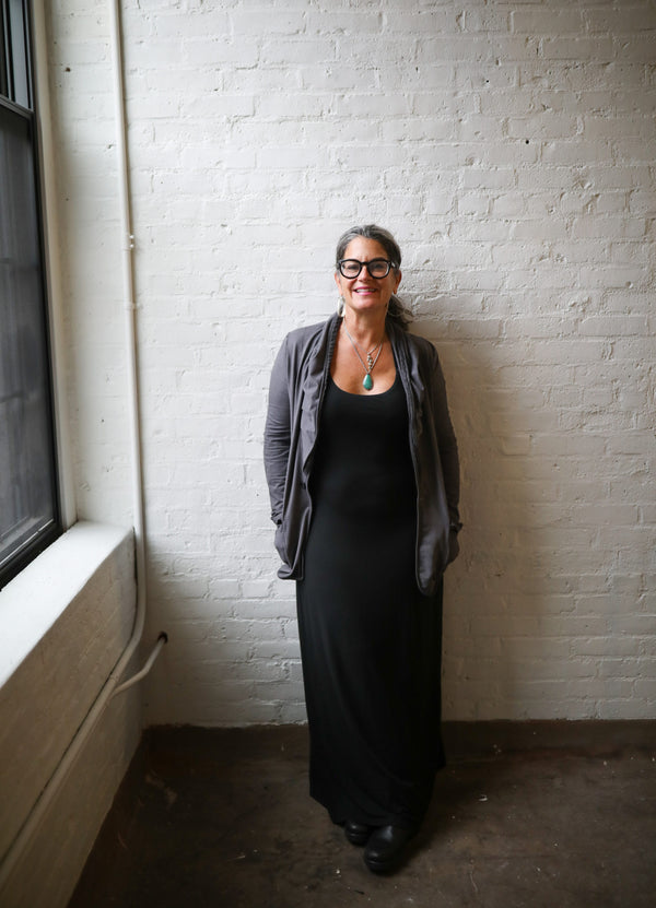 Woman in a long dark dress, bold glasses and hair pulled back, smiling, her back against wall of distressed brick.