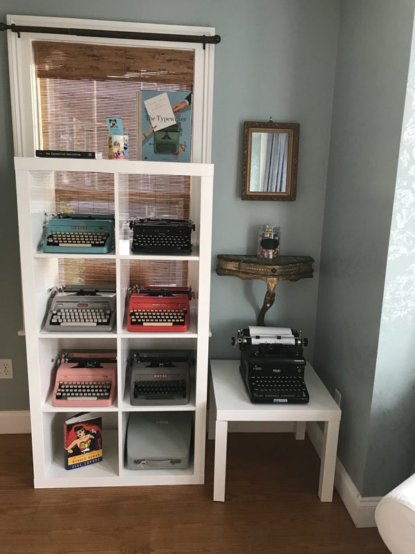 The corner of a room, a tall shelf filled with typewriters, a small table with another typewriter, and a small mirror hanging above.