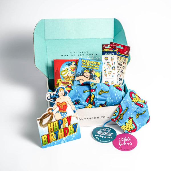 An open gift box overflowing with wonder woman gifts including stickers, notecards and face masks.
